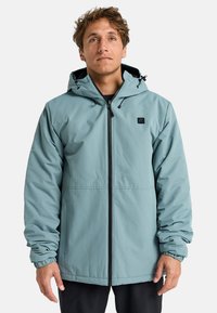 Chaqueta impermeable de color azul claro con capucha, cremallera frontal, puños elásticos y un pequeño logo negro en el pecho. Textura suave, diseño casual.