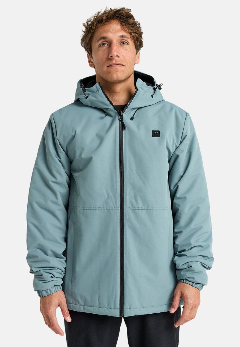 Chaqueta impermeable de color azul claro con capucha, cremallera frontal, puños elásticos y un pequeño logo negro en el pecho. Textura suave, diseño casual.