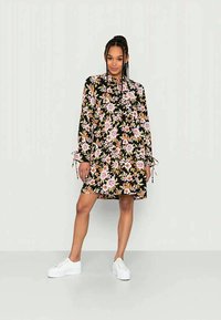 Vestido floral com mangas longas, fundo preto, flores rosas e laranjas, corpete franzido e punhos com cordão, combinado com sapatos brancos.