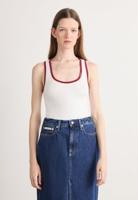 Débardeur blanc à côtes avec des bretelles rouge foncé, associé à une jupe en denim bleue taille haute avec une étiquette de logo et des boutons avant.
