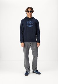Marineblauwe hoodie met groot boomlogo in blauw, voorzak, ribbelboordjes, gecombineerd met grijze broek en blauwe sneakers met drie strepen.