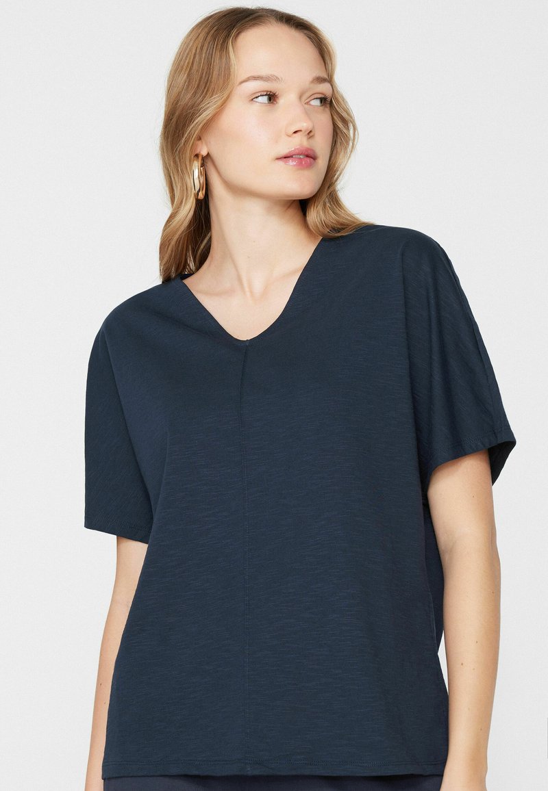 Femme aux cheveux clairs et boucles d'oreilles créoles dorées, portant un haut bleu marine à manches courtes et col en V, regardant vers sa droite sur un fond uni.