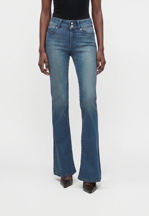 Femme portant un jean évasé taille haute bleu et des chaussures noires à bouts pointus, debout devant un fond blanc uni.