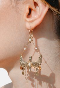 Boucles d'oreilles en métal doré en forme d'oiseau avec des détails complexes, chaînes suspendues et perles décoratives blanches et irisées, présentées sur une oreille.