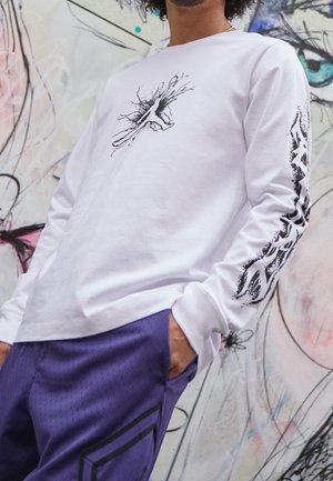 White long-sleeve shirt with black graphic design featuring abstract lines. Paired with purple perforated shorts and side pockets.
