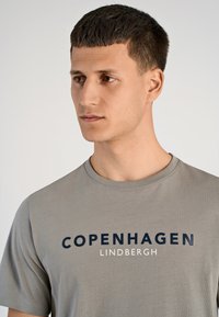 Grijze katoenen t-shirt met ronde hals, korte mouwen en dikke marineblauwe tekst: "KOPENHAGEN" en "LINDBERGH" op de voorkant.