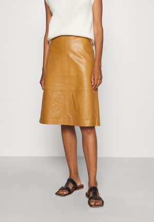 Mujer con un top sin mangas de punto blanco, falda de cuero color marrón claro hasta la rodilla y sandalias planas negras con tiras anchas.