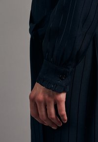 Handen vilar vid sidan i en mörk randig långärmad klädsel med knäppta manschetter och volangdetaljer.