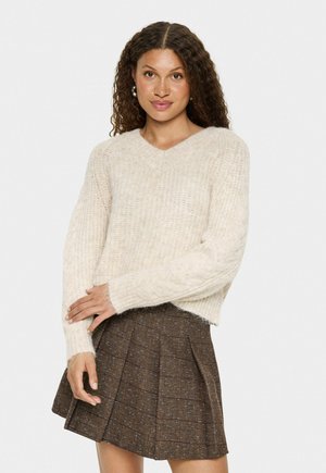 Cremfarbener, grob gestrickter Pullover mit V-Ausschnitt, langen Ärmeln und gerippten Bündchen, kombiniert mit einem braunen karierter Faltenrock. Strukturiertes und gestuftes Design.