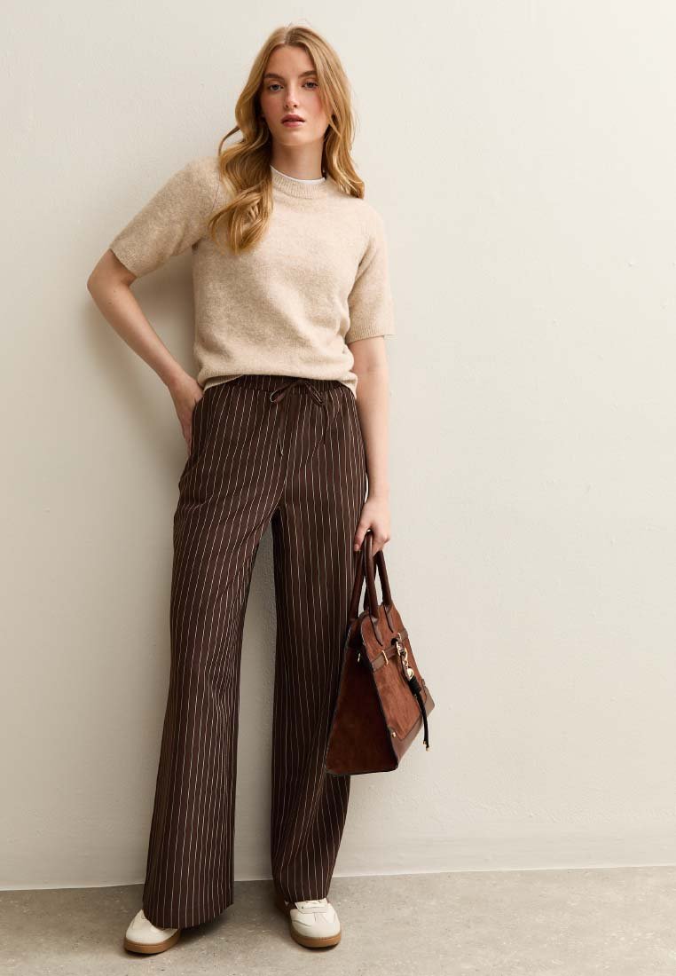 Beige short-sleeve sweater, brown striped wide-leg pants with drawstring. Brown handbag with metal accents, white sneakers. Plain background.