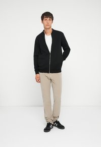Sort zip-up sweatshirt med ribbede manchetter, parret med lyse beige bukser og sorte sneakers. Modellen står foran en ensfarvet baggrund.