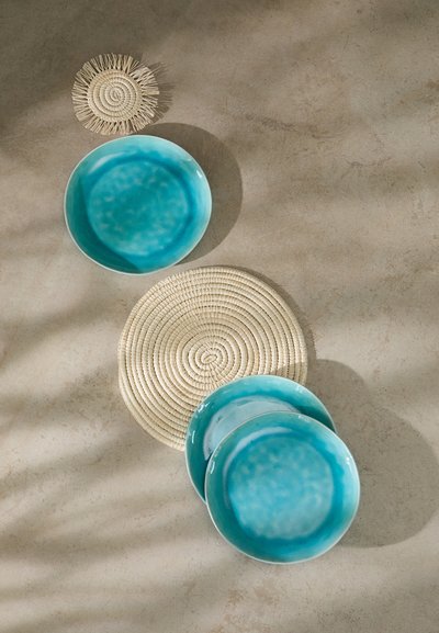 Set of three turquoise ceramic plates stacked beside a round, natural woven placemat with a spiral design, on a light textured surface.
