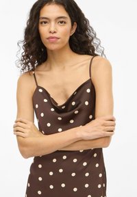 Rjava obleka z ovratnikom v obliki kapuce, okrašena z belimi pikami, zanje tanke naramnice in svilnato teksturo, ki mehko pada čez telo.