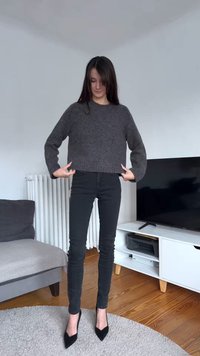 Siv skrajšan pulover z dolgimi rokavi in neravnim robom, kombiniran s črnimi oprijetimi jeans hlačami in črnimi čevlji z zašiljenim prstom. Lahka tekstura, sproščen kroj.