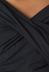 Lähikuva tummasta kankaasta, joka on kiertynyt ristikkäin vaaleanruskealla iholla, viitaten vaatteen kaula-aukon tai kääretakaan yksityiskohtiin.