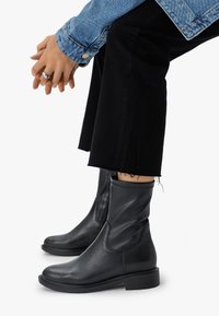 Mustat nahkasaappaat nilkkapituiset, joissa on sileä pinta, joustavat sivut ja litteä pohja; yhdistetty mustiin rispaantuneisiin housuihin ja farkkutakkiin.