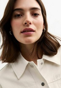 Chemise blanche à boutons avec un col structuré. Dotée de quincaillerie de boutons en argent. Le modèle a les cheveux bruns mi-longs et un maquillage naturel.