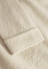 Krémová vlněná tkanina s měkkým, strukturovaným povrchem a podhnutým lemem. Materiál vypadá teple a útulně.