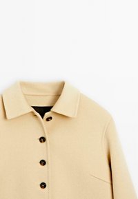 Beige wollen jas met een kraag, voorzien van vijf donkere knopen, een borstas, en een zachte, gestructureerde oppervlakte.