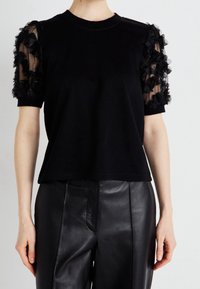 Musta T-paita, jonka lyhyet hihat ovat läpikuultavaa kangasta ja joissa on kukkakuvioita. Pään osa on kiinteää mustaa puuvillaa. Yhdistetty mustien nahkahousujen kanssa.