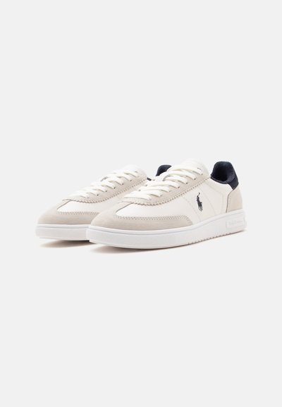Ζευγάρι λευκά και μπεζ sneakers χαμηλού προφίλ με κορδόνια μπροστά, μαύρη ταμπέλα στη φτέρνα και μικρό μαύρο λογότυπο στο πλάι.