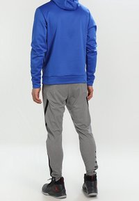 Blaue Kapuzenjacke mit glatter Textur, graue tapered Hose mit schwarzen Seitenelementen und schwarze Sneaker mit roten Akzenten.