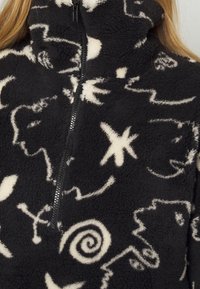Pullover in pile nero con collo alto, caratterizzato da motivi astratti bianchi. Chiusura con zip sul davanti e una finitura morbida e testurizzata.