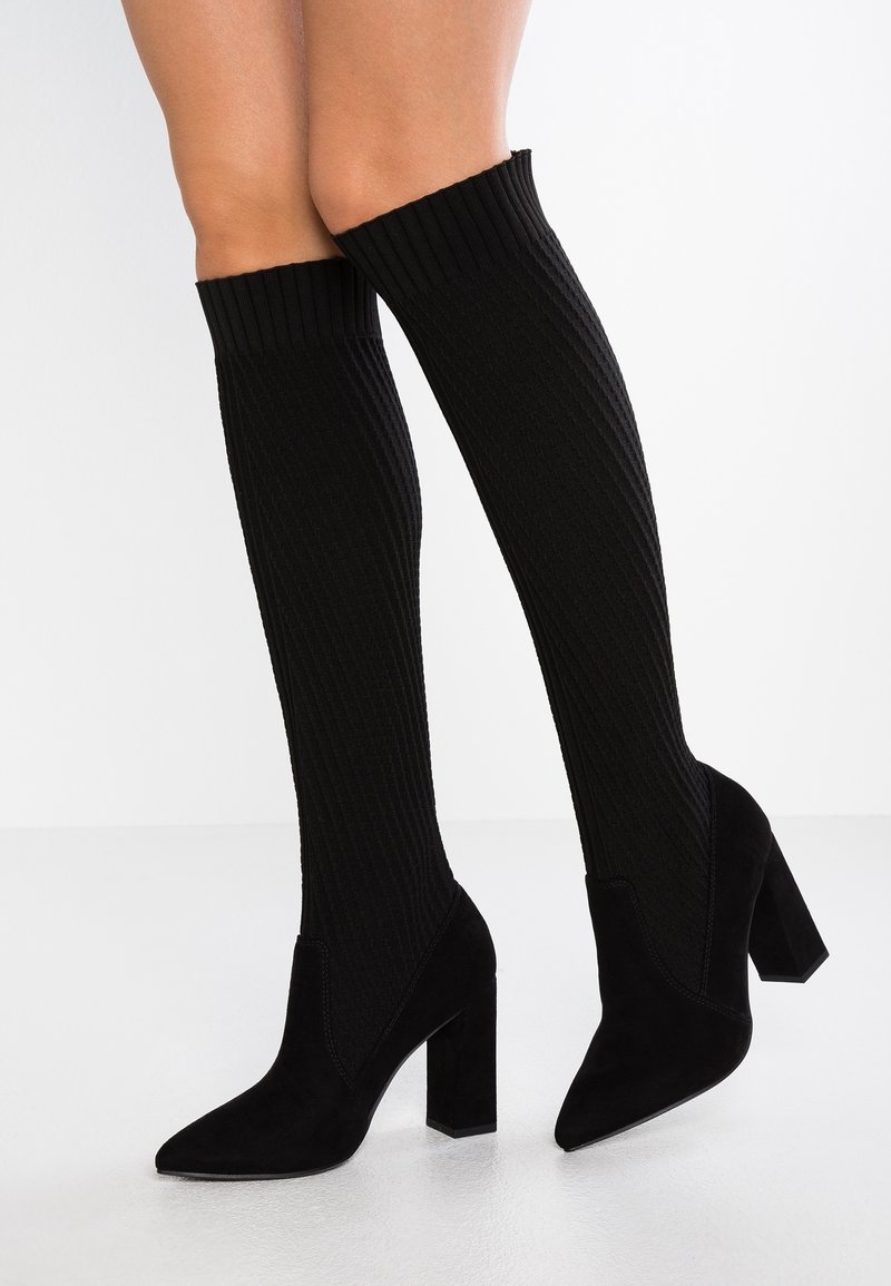Bottes chaussettes noires hauteur genou à côtes, avec bouts pointus et talons blocs, portées sur jambes nues sur un fond clair uni.