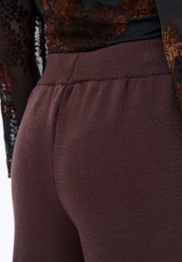 Bordeaux stretchbroek met een zachte textuur en elastische tailleband, zijwaarts weergegeven, waarbij de getailleerde silhoët en het materiaal worden benadrukt.