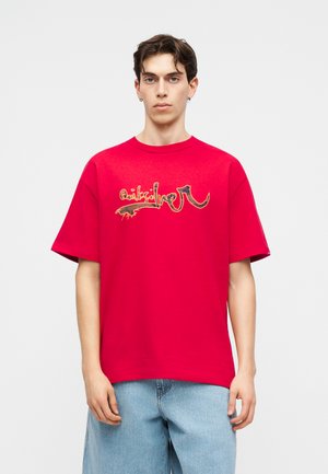 Joven con una camiseta roja de talla grande con diseño gráfico abstracto y pantalones cortos vaqueros azul claro, de pie sobre un fondo liso.