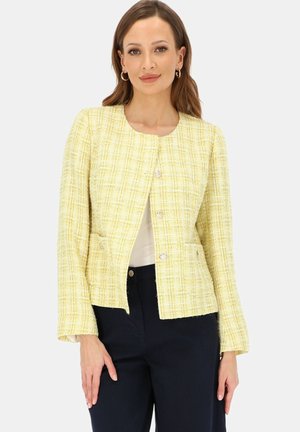 Kobieta ubrana w jasną żółtą, w kratę, zapinaną na guziki kurtkę z długimi rękawami i czarne spodnie, stojąca na jednolitym tle.