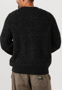 Man in een zwart chunky gebreide trui en beige corduroy broek met een groen label "Workwear Inspired Apparel" op de achterzak.