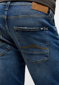 Blå denimjeans med framträdande sömmar och två bakfickor. En läderetikett med "MUSTANG" är synlig på midjebandet.