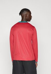 Κόκκινο πουκάμισο με μακριά μανίκια, σχέδιο με κυματιστή γραμμή και λεπτομέρεια στο γιακά σε τυρκουάζ. Ελαφρύ υλικό, εφαρμοστή γραμμή και στρογγυλή λαιμόκοψη.