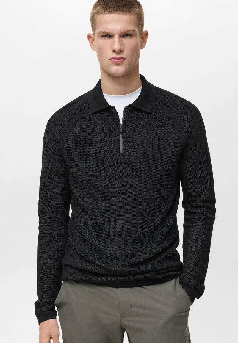 Pullover nero a maglia con colletto con zip e maniche raglan, caratterizzato da un tessuto testurizzato e un design aderente. Indossato sopra una camicia bianca.