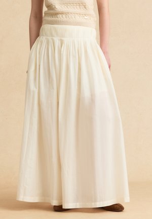 Donna che indossa una gonna lunga, bianca, arricciata con tasche, abbinata a un top beige testurizzato senza maniche e scarpe marroni.