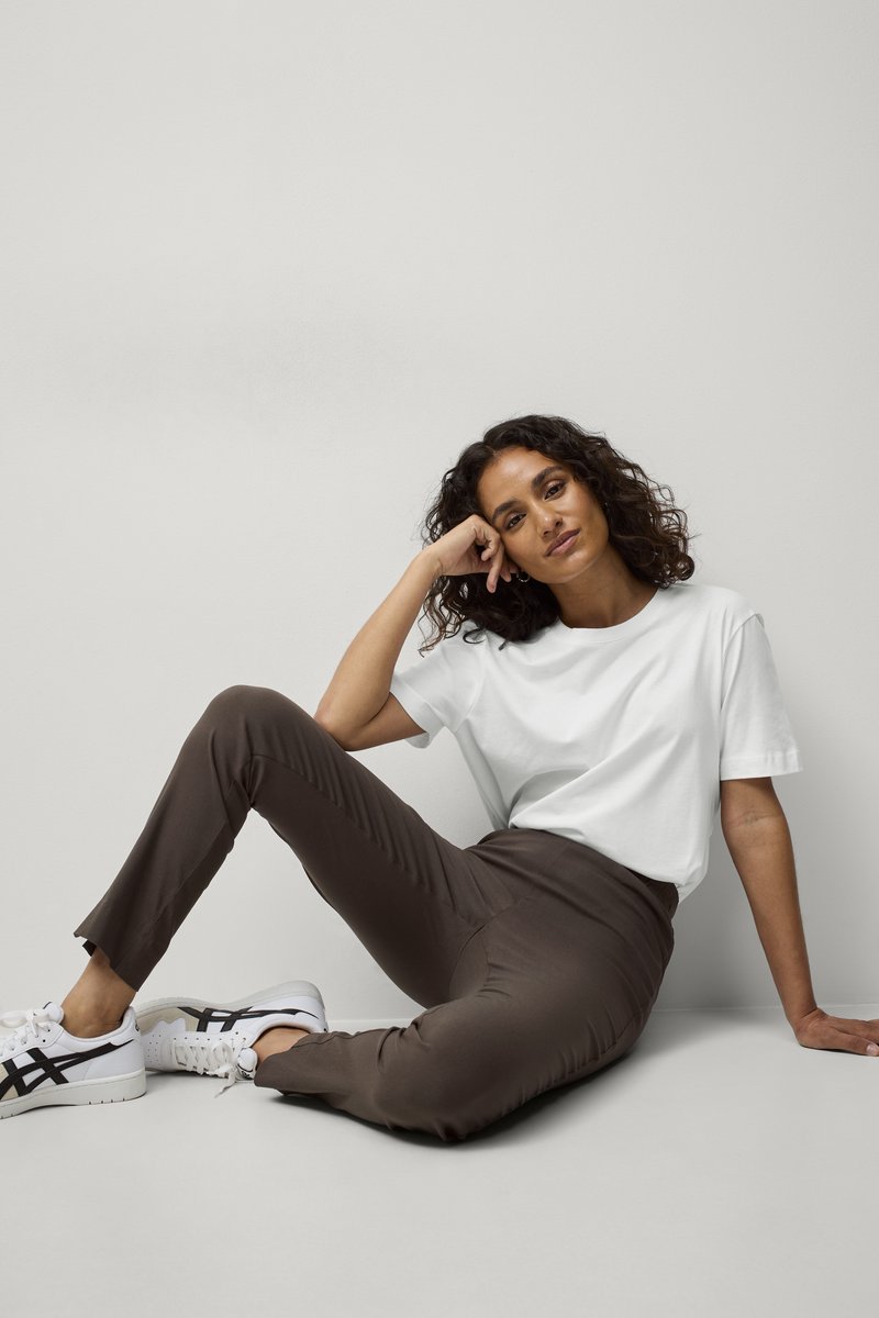 T-shirt en coton blanc associé à un pantalon élastique marron et des baskets blanches avec des accents noirs. Modèle assis, présentant une tenue décontractée.