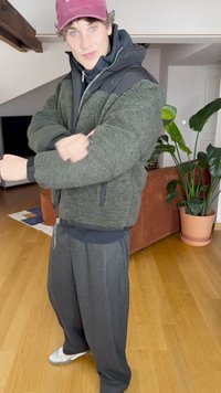 Olijfgroene fleecejas met een zwarte capuchon, een bulky ontwerp en ritszakken, gecombineerd met losse grijze broek en witte sneakers.