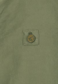 Oliegrøn stof med en struktureret overflade, der har en firkantet lap med et rundt, metallisk logo fastgjort i midten.