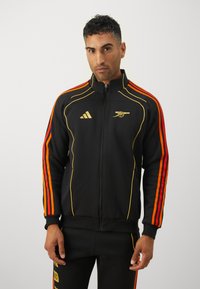 Veste noire zippée avec des manches rayées orange et rouge, présentant des accents dorés et un logo. Fabriquée dans un matériau lisse avec une coupe ajustée.