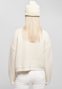 En blond person vänd bort från kameran, iklädd en krämfärgad stickad hjälm, en tjock tröja och vita byxor, framför en enfärgad grå bakgrund.