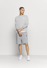 Completo grigio con felpa e pantaloni corti, entrambi realizzati in tessuto morbido. Presenta un logo Nike bianco sul petto e sui pantaloni corti. Indossato con sneakers bianche.