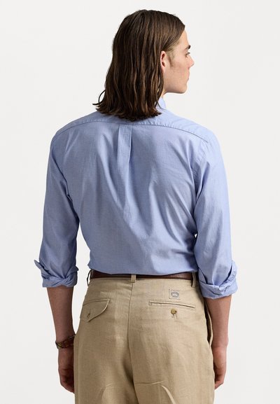 Homem com cabelo castanho até aos ombros, vestindo uma camisa de mangas compridas azul clara com as mangas arregaçadas e calças beiges, visto por trás.