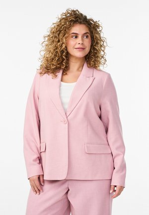 Kobieta o kręconych włosach nosi jasnoróżową marynarkę i dopasowane spodnie na białej bluzce, patrząc w lewo na jednolitym tle.