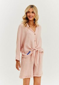 Svetlé ružové ľanové tričko s viazaním vpredu a gombíkmi, v kombinácii s ladiacimi ľanovými šortkami s roll-up lemami. Viditeľná jemná textúra.