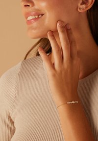 Bracelet en or avec de petites perles et des accents en forme de cœur, porté sur une main avec un vernis à ongles neutre, aux côtés d'un top beige côtelé.