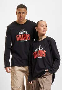 Zwarte langmouwshirts hebben een bold "HELVETIC GUARDS" ontwerp in rood en wit. Het materiaal is katoen met een relaxte pasvorm, gecombineerd met beige broeken.