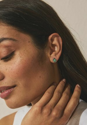 Woman with closed eyes touching her neck, wearing a silver stud earring with a green stone, against a neutral background.