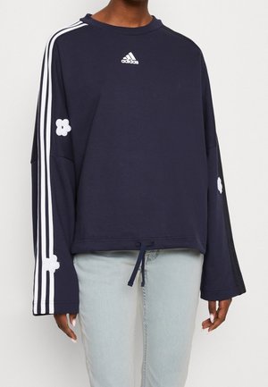 Granatowa bluza Adidas z białymi paskami i naszywkami w kształcie kwiatów na rękawach, noszona z jasnoniebieskimi dżinsami, głowa osoby niewidoczna.