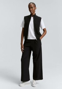 Schwarzes geripptes Shirt mit hohem Kragen und Vorderreißverschluss, getragen über einem weißen T-Shirt, kombiniert mit weiten schwarzen Hosen und weißen Turnschuhen.